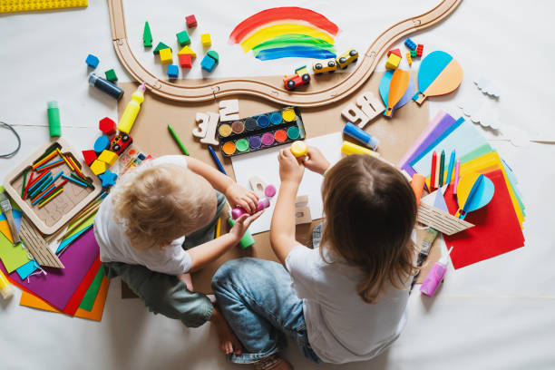 Two children playing with wooden toys, art supplies, and loose parts spread across the floor