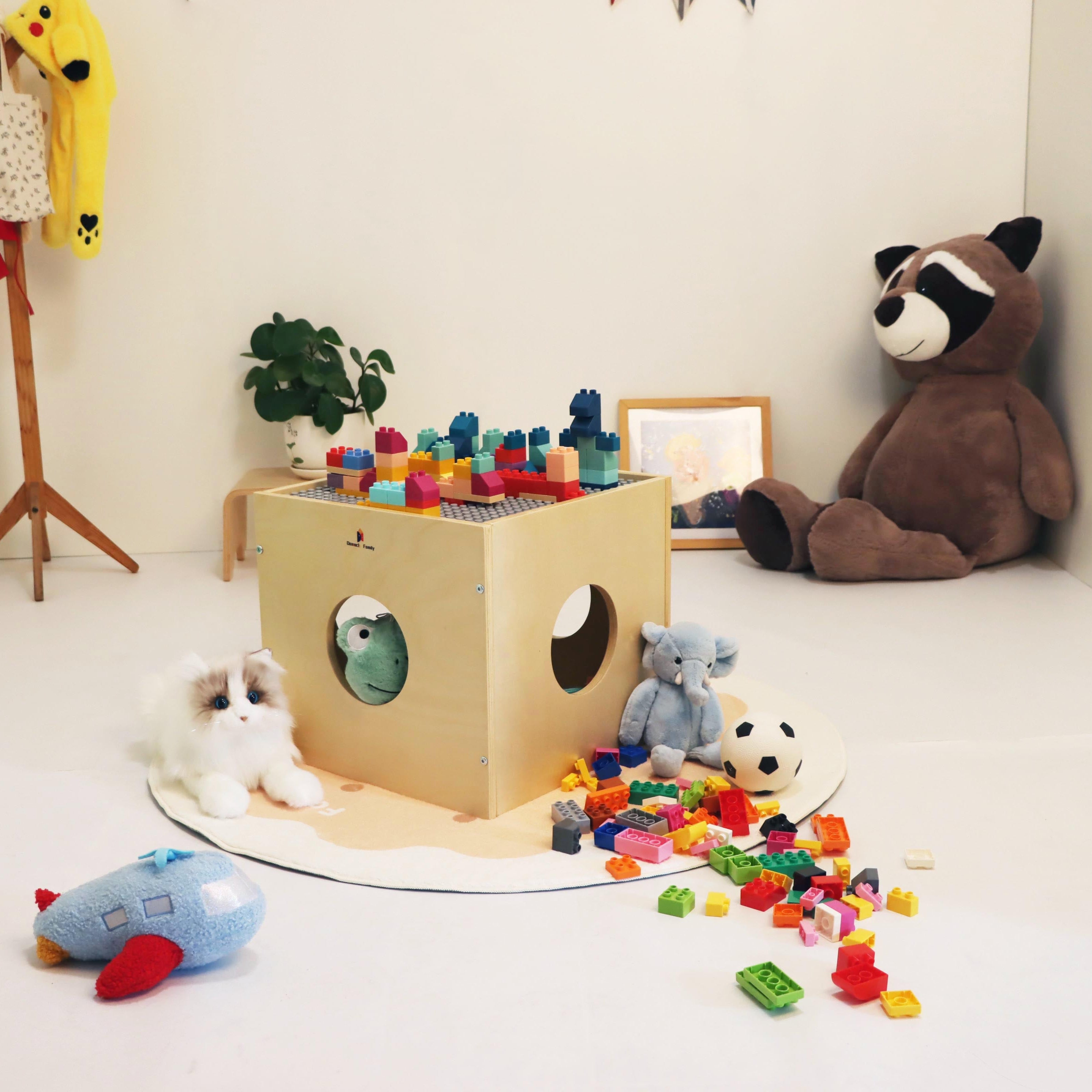 Children's play area with a cardboard box toy, colorful blocks, and stuffed animals on a white floor.