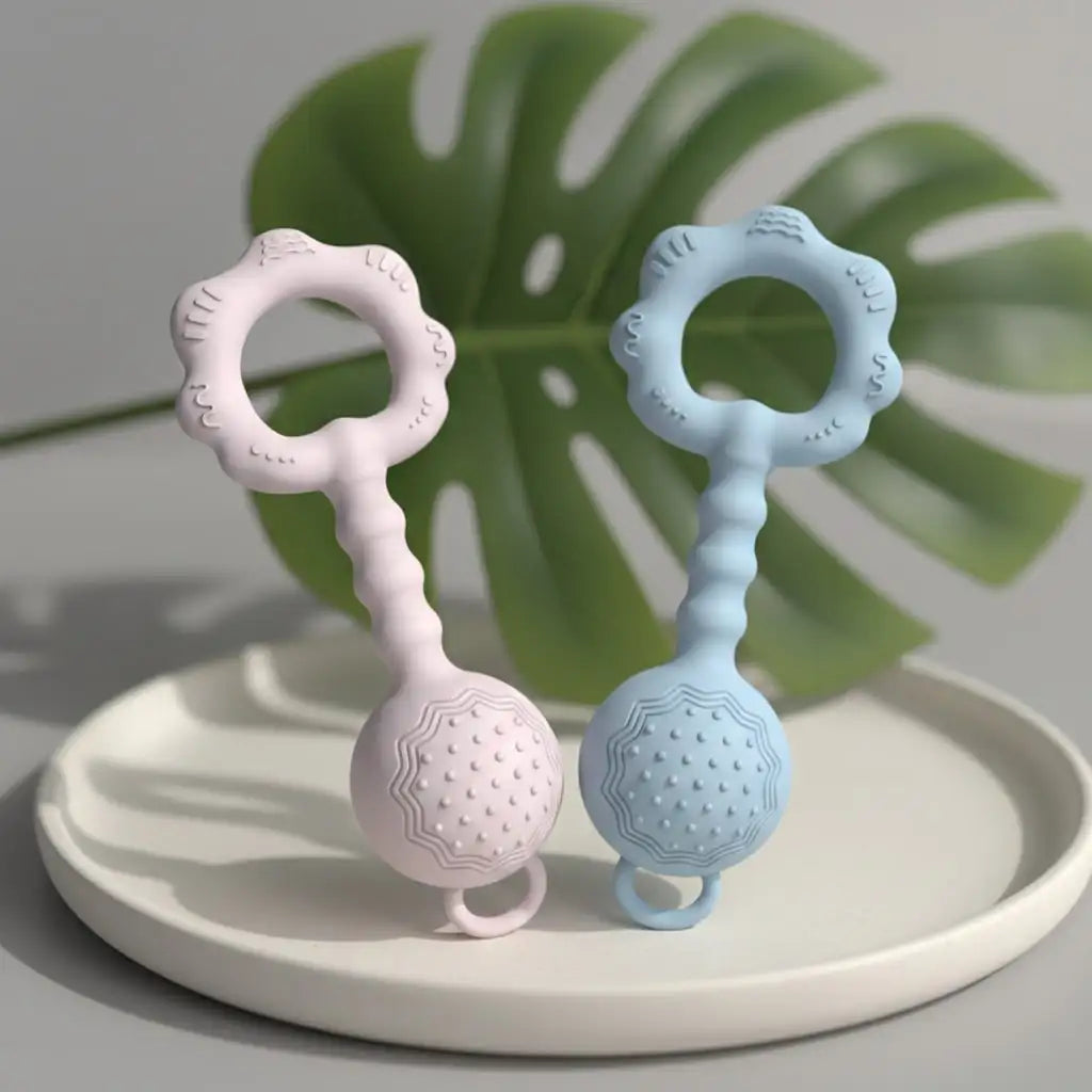 Two silicone baby teething toys, one light pink and one light blue, standing upright on a white ceramic tray with a blurred green monstera leaf in the background.