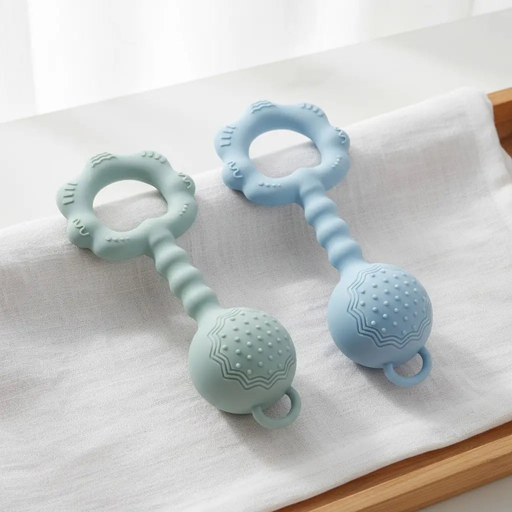 Two silicone baby teething toys with textured ball ends and flower-shaped rings, shown in mint green and light blue on a white fabric surface.
