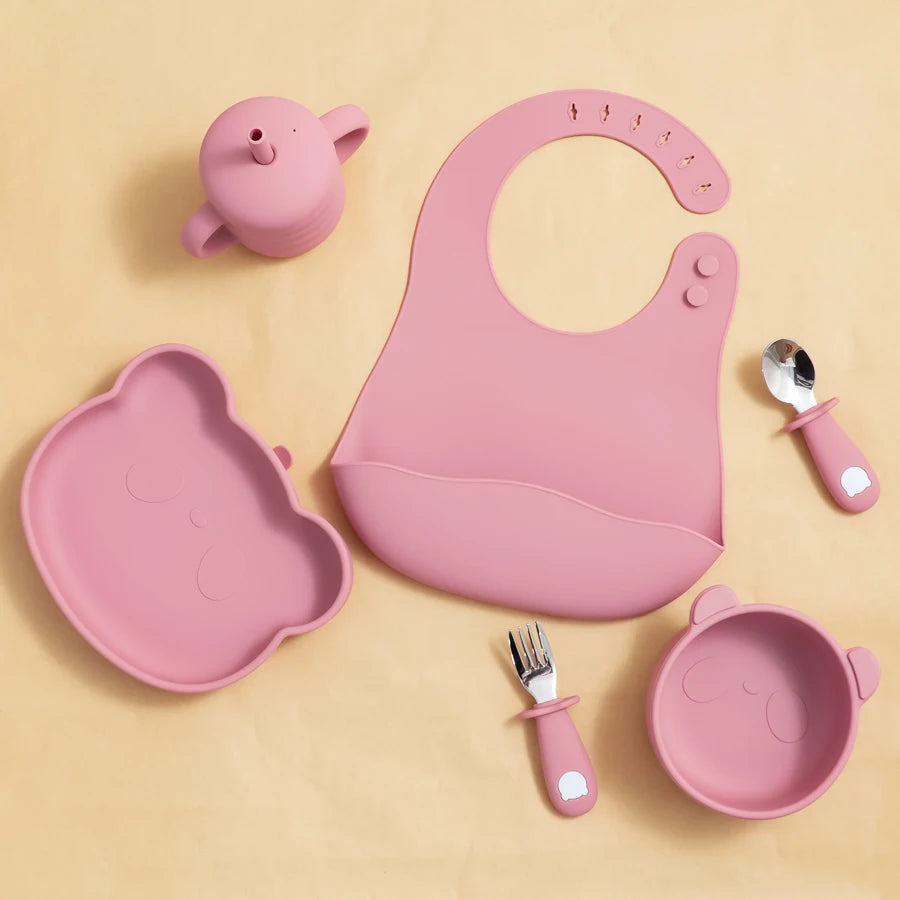 Pink children's dining set including a bib, plate, cup, bowl, and utensils on a beige background.