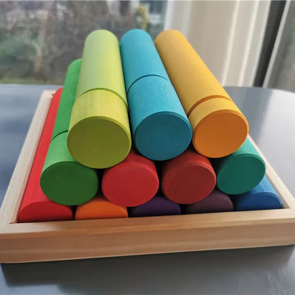 A set of colorful wooden cylinder blocks arranged in a light wood tray on a tabletop near a window.