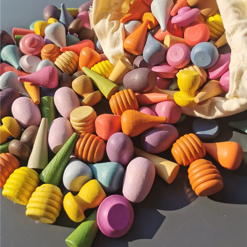 A pile of colorful wooden toy shapes and pegs spilling out of a cream fabric bag onto a dark surface. The toys include cones, ribbed honeycomb-like pieces, rounded blocks, and pegs in many pastel and bright colors such as pink, orange, yellow, green, blue, purple, and brown.