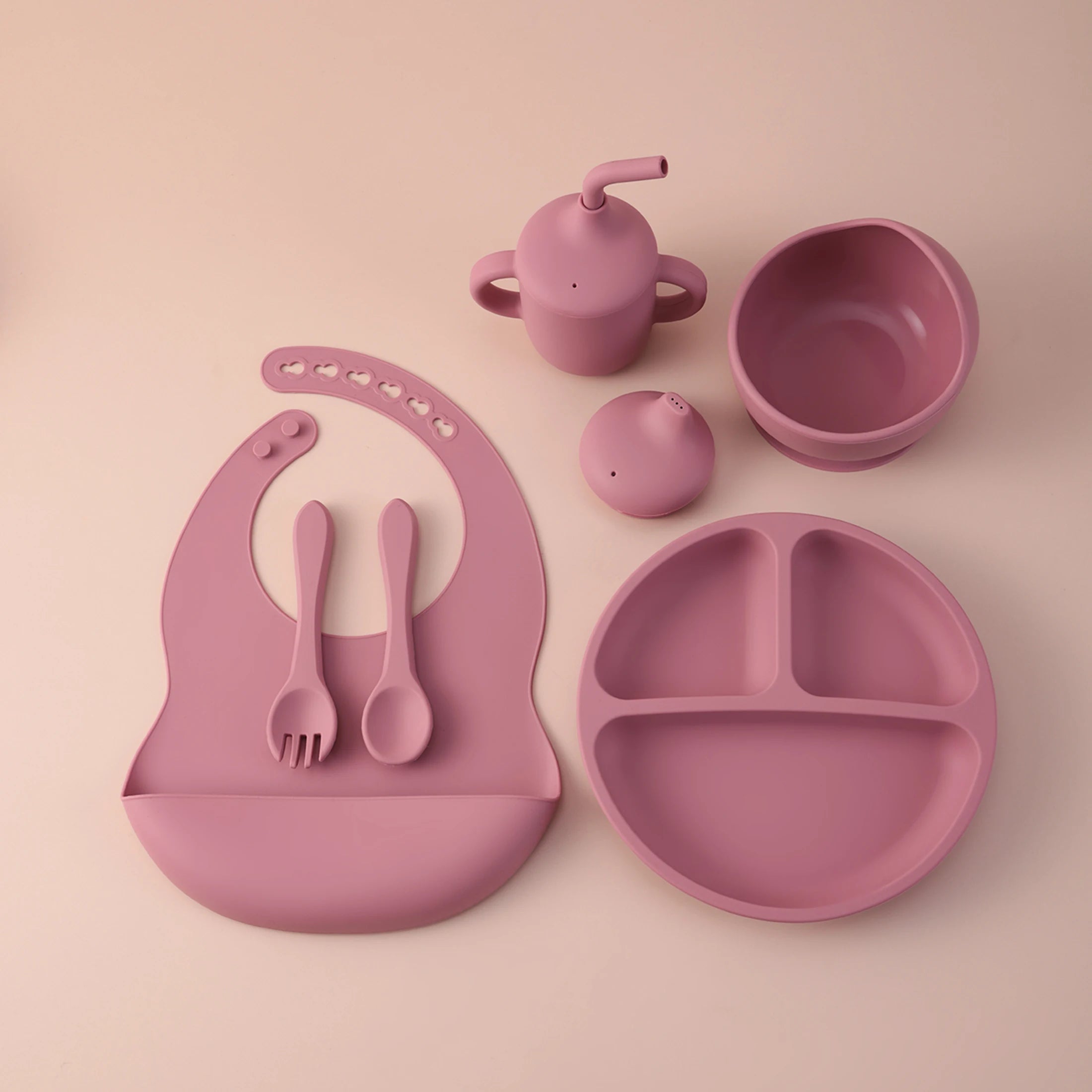 A coordinated baby feeding set in a muted dusty pink color, including a silicone bib with integrated utensils, a divided plate, a small handled tumbler, a small bottle, and a bowl, arranged on a plain warm beige background.