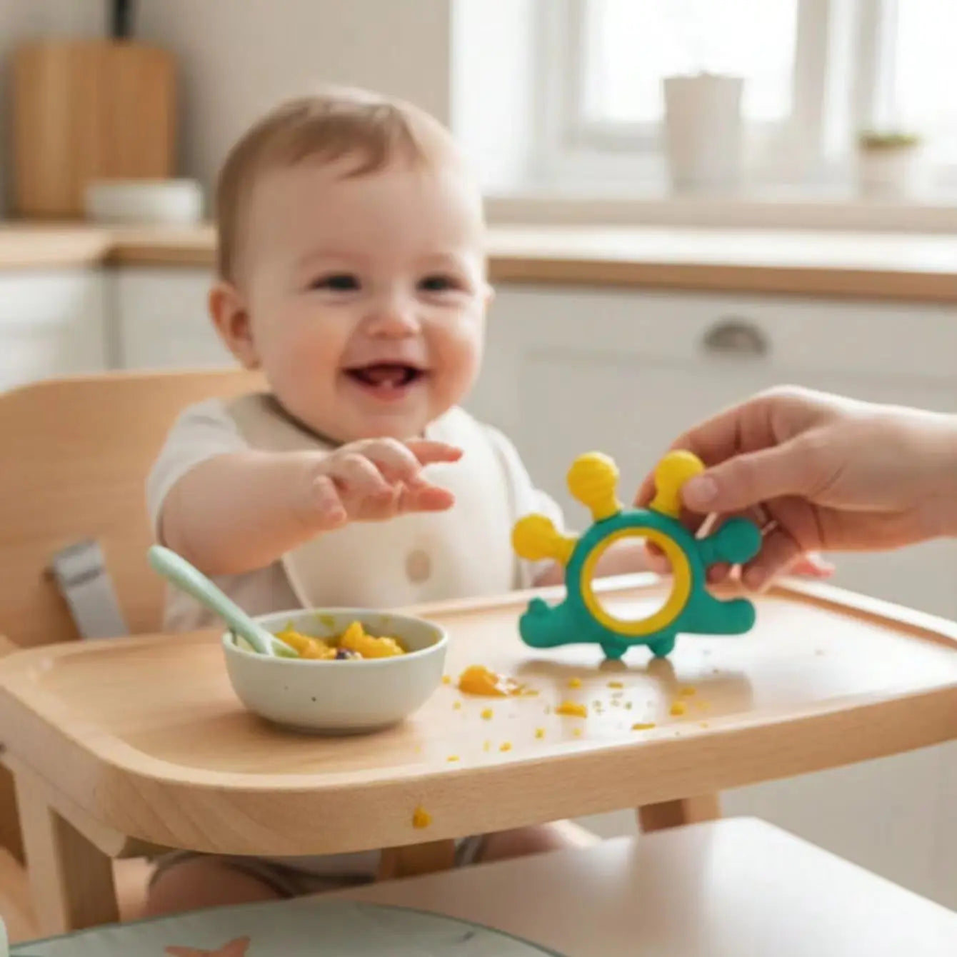 A baby sitting in a wooden high chair smiling while reaching for a green and yellow teething toy held by an adult hand. A small bowl of food and a spoon sit on the tray in a bright, softly blurred kitchen setting.