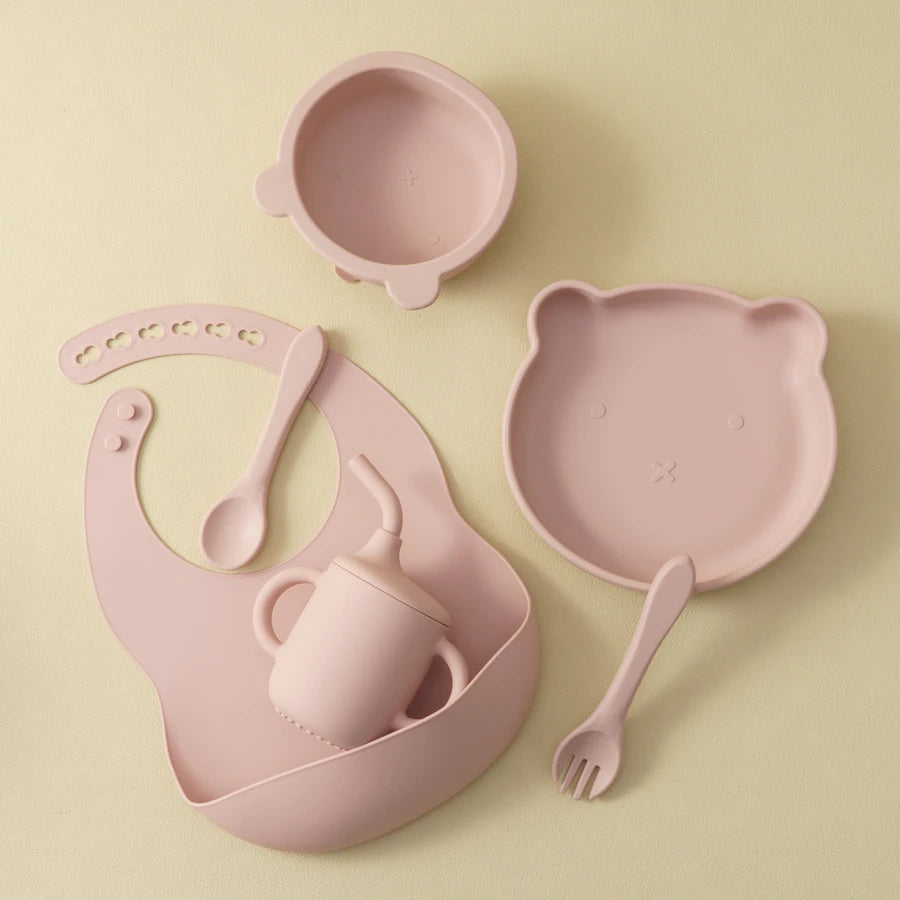 Pink children's dining set including bowl, plate, cup, spoon, and fork on a beige background