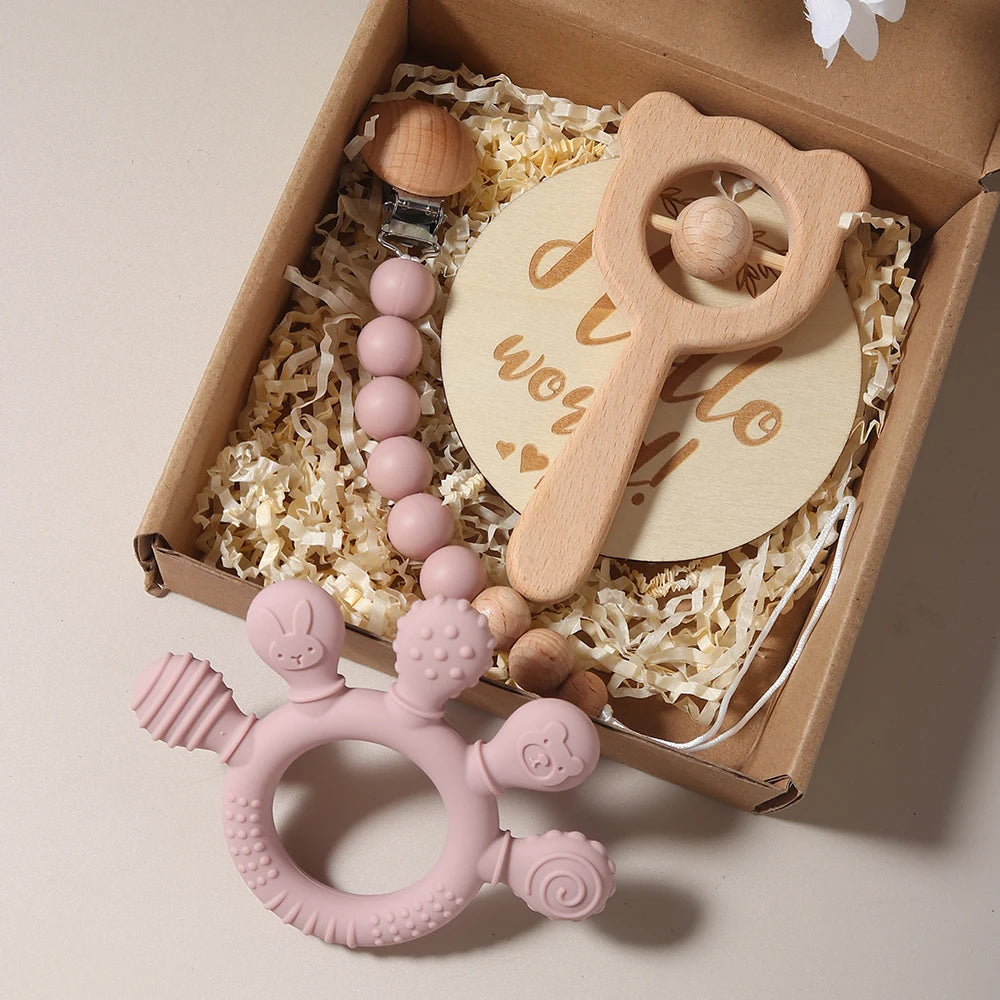 Set of baby teething toys including a pink silicone teether, wooden rattle, and pacifier clip in a box.