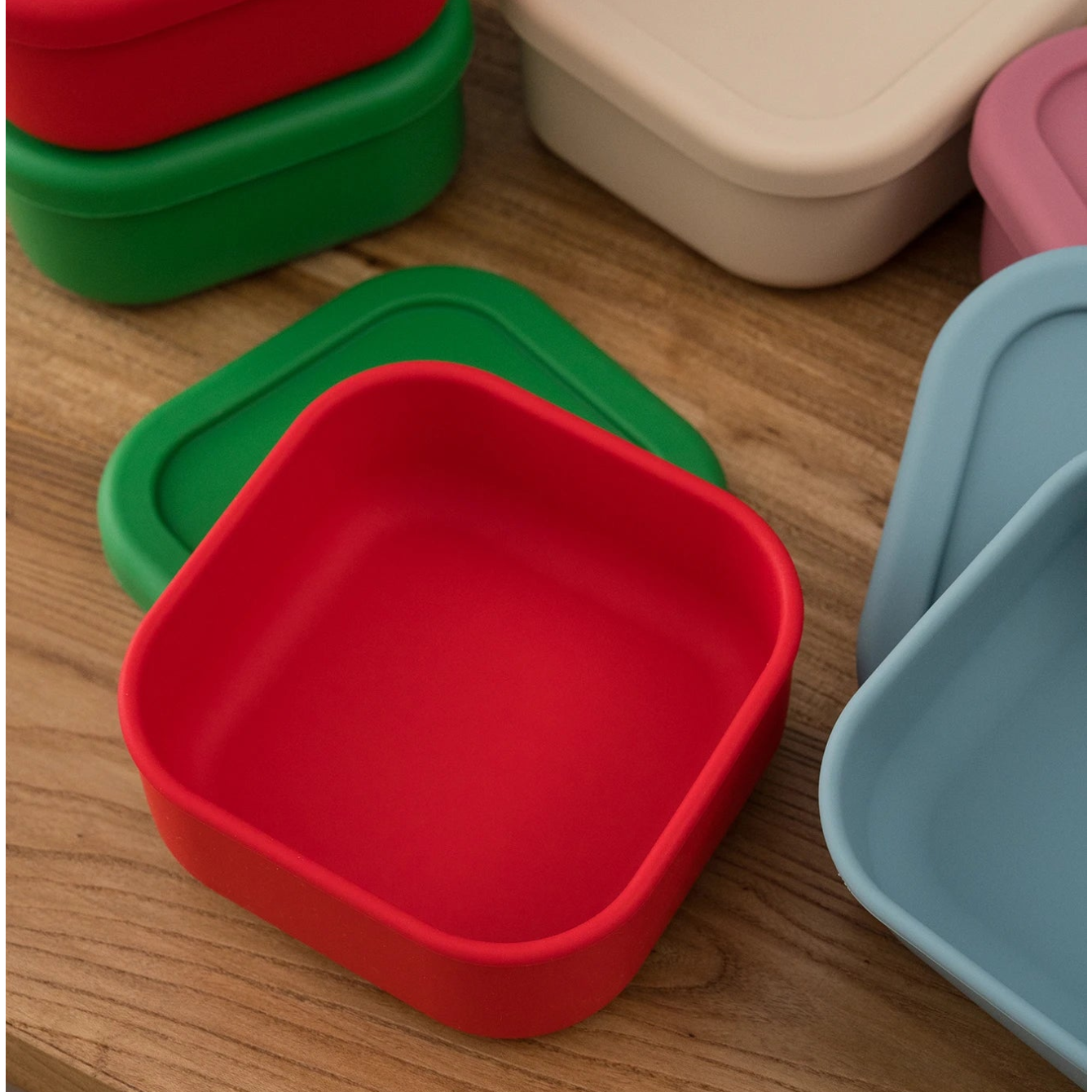 Red square silicone snack container with lid beside colorful containers