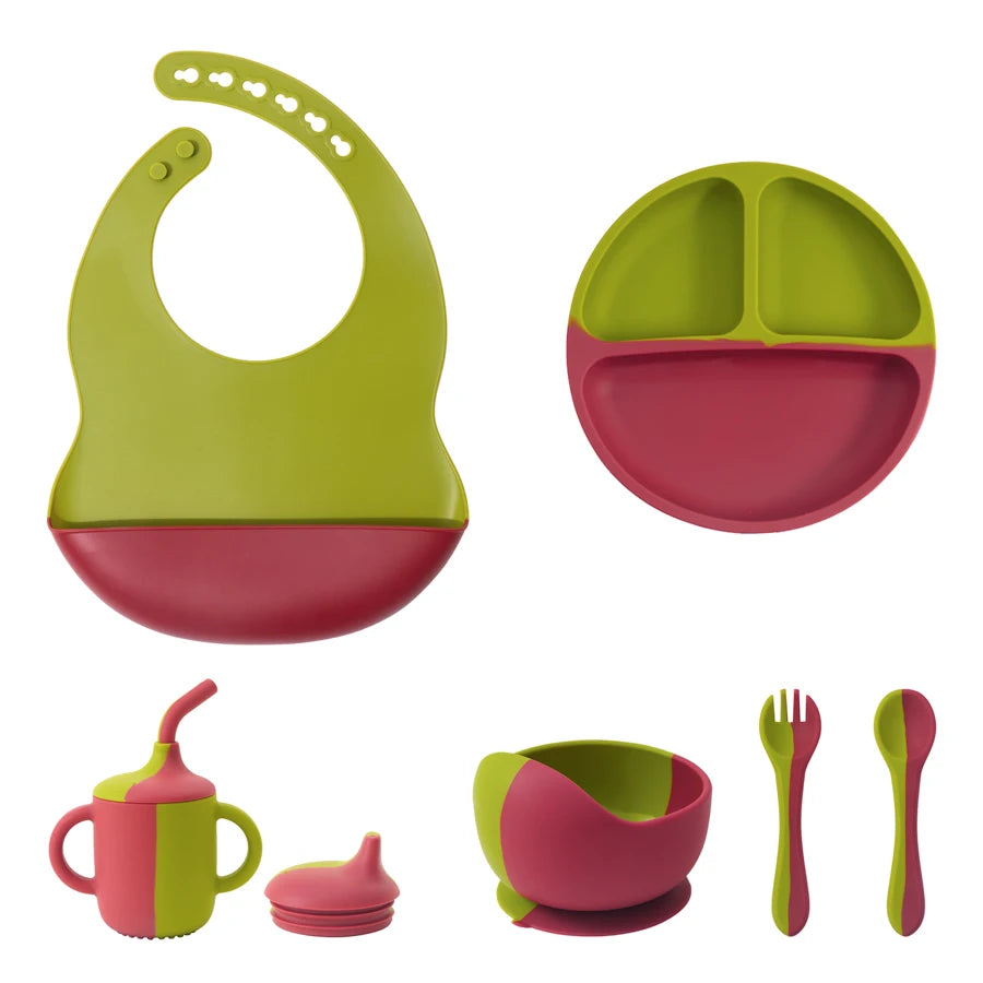 A baby feeding set with multiple silicone pieces in lime green and pink, including a bib, divided plate, sippy cup with lid, bowl, fork, and spoon, shown on a plain white background.