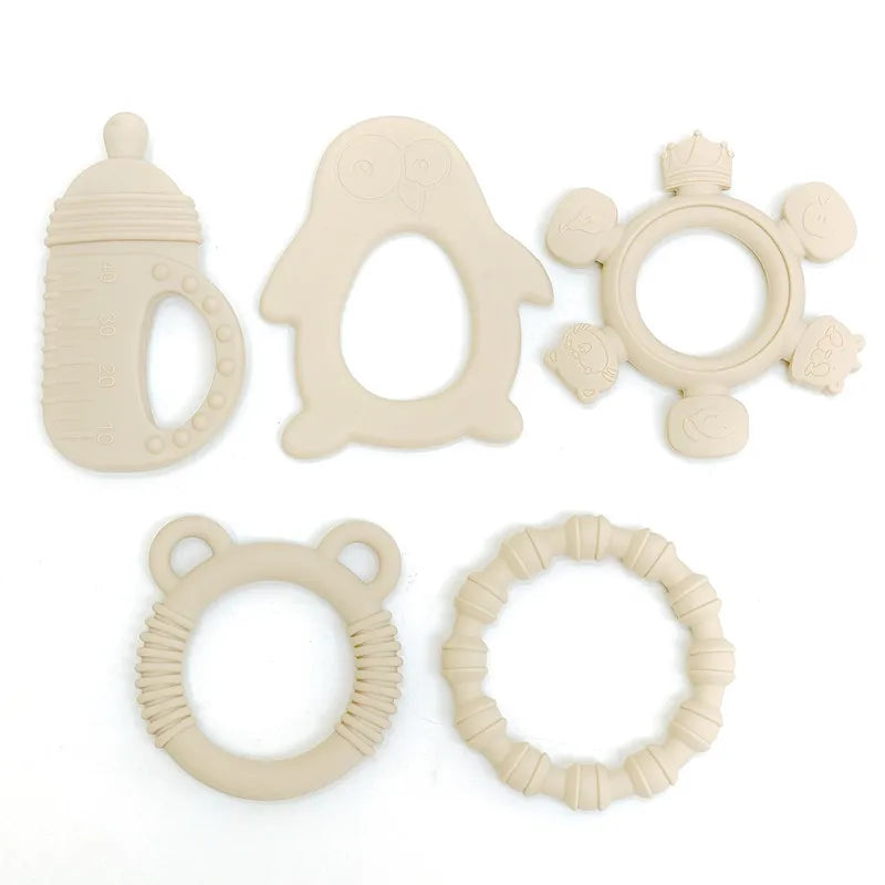 Five beige silicone baby teething toys arranged on a plain white background, including a bottle-shaped toy, an owl teether, a flower ring with animal details, a bear ear ring, and a textured circular ring.