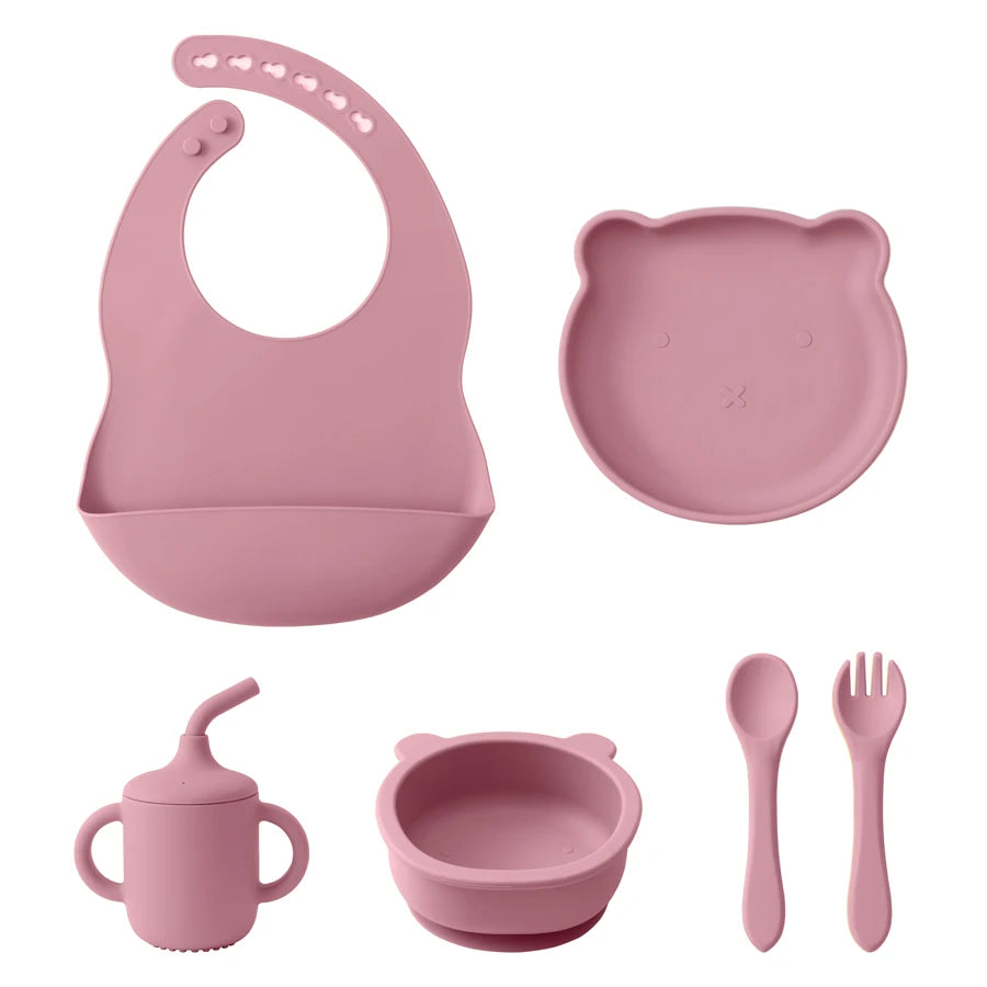 Pink baby dining set with bib, plate, cup, bowl, spoon, and fork on a white background