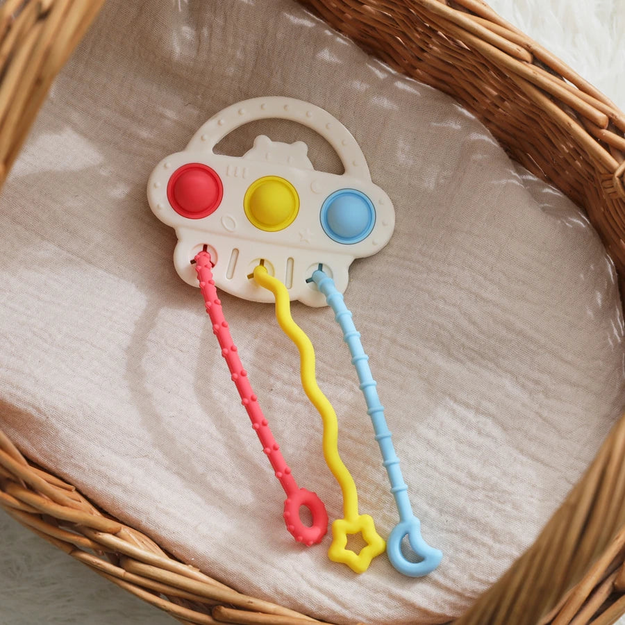 A baby sensory toy with a white cloud-shaped handle and three colorful plastic loops (red, yellow, and blue) resting on a beige fabric liner inside a wicker basket.