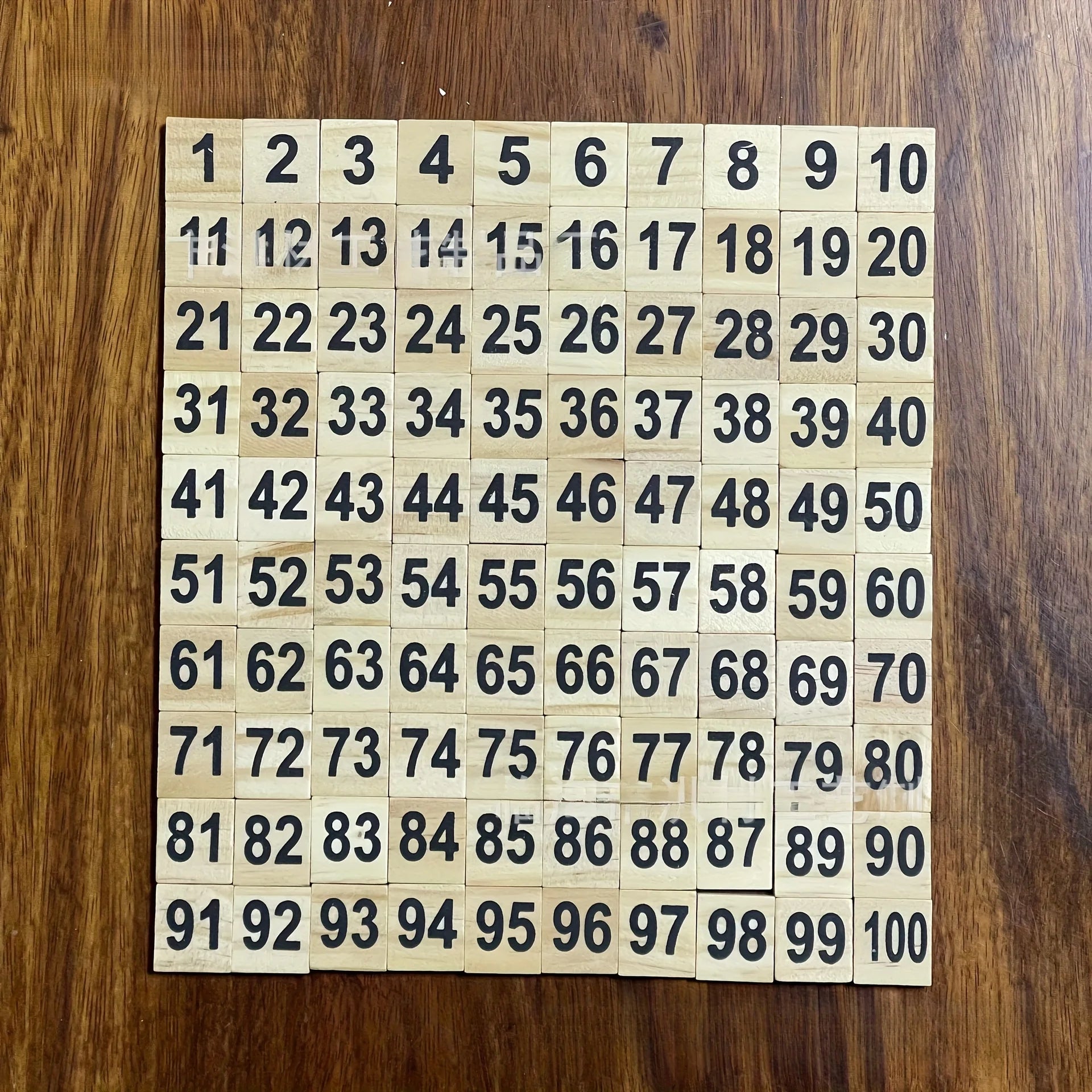 A set of wooden number tiles arranged in a grid on a dark wood surface, showing numbers from 1 to 100 in black print.