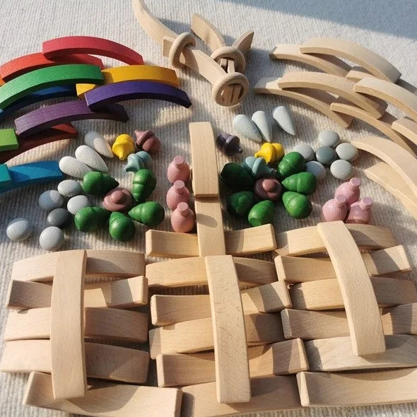 A wooden toy construction set arranged on a light fabric background, including natural wood blocks, curved wooden beams, colorful wooden hoops, small painted wooden pieces, and loose stones.