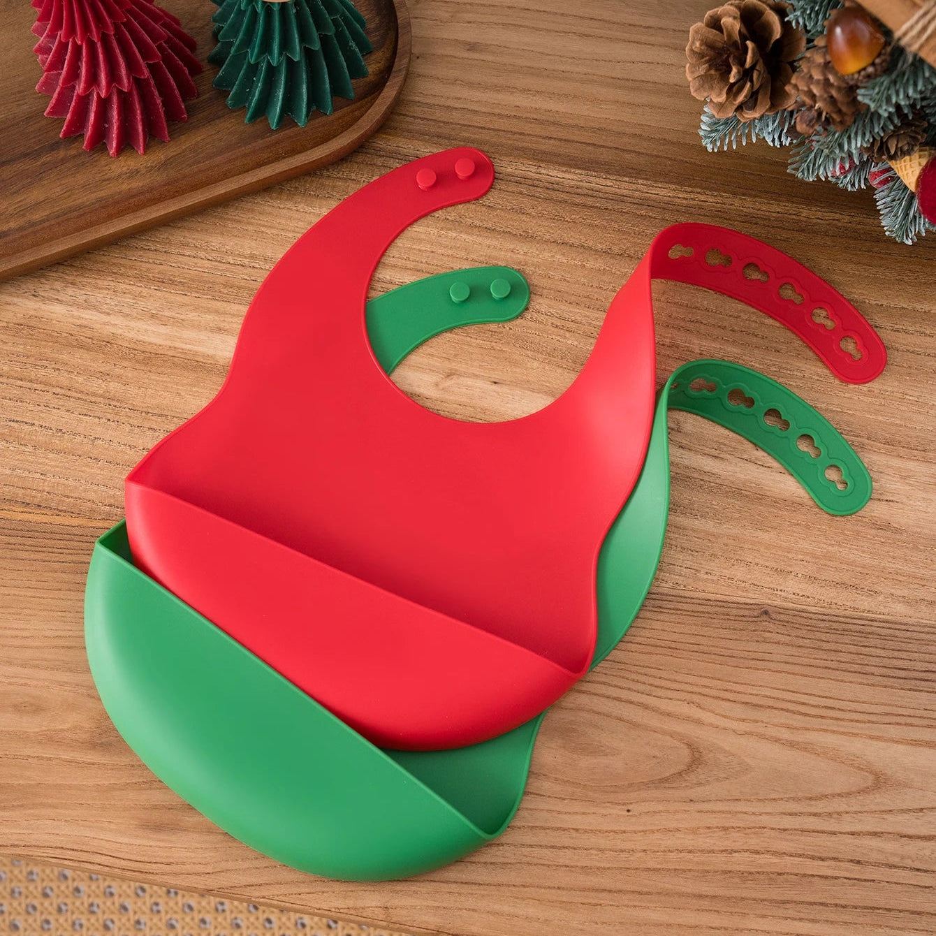 Red and green silicone bibs on a wooden table with holiday decor