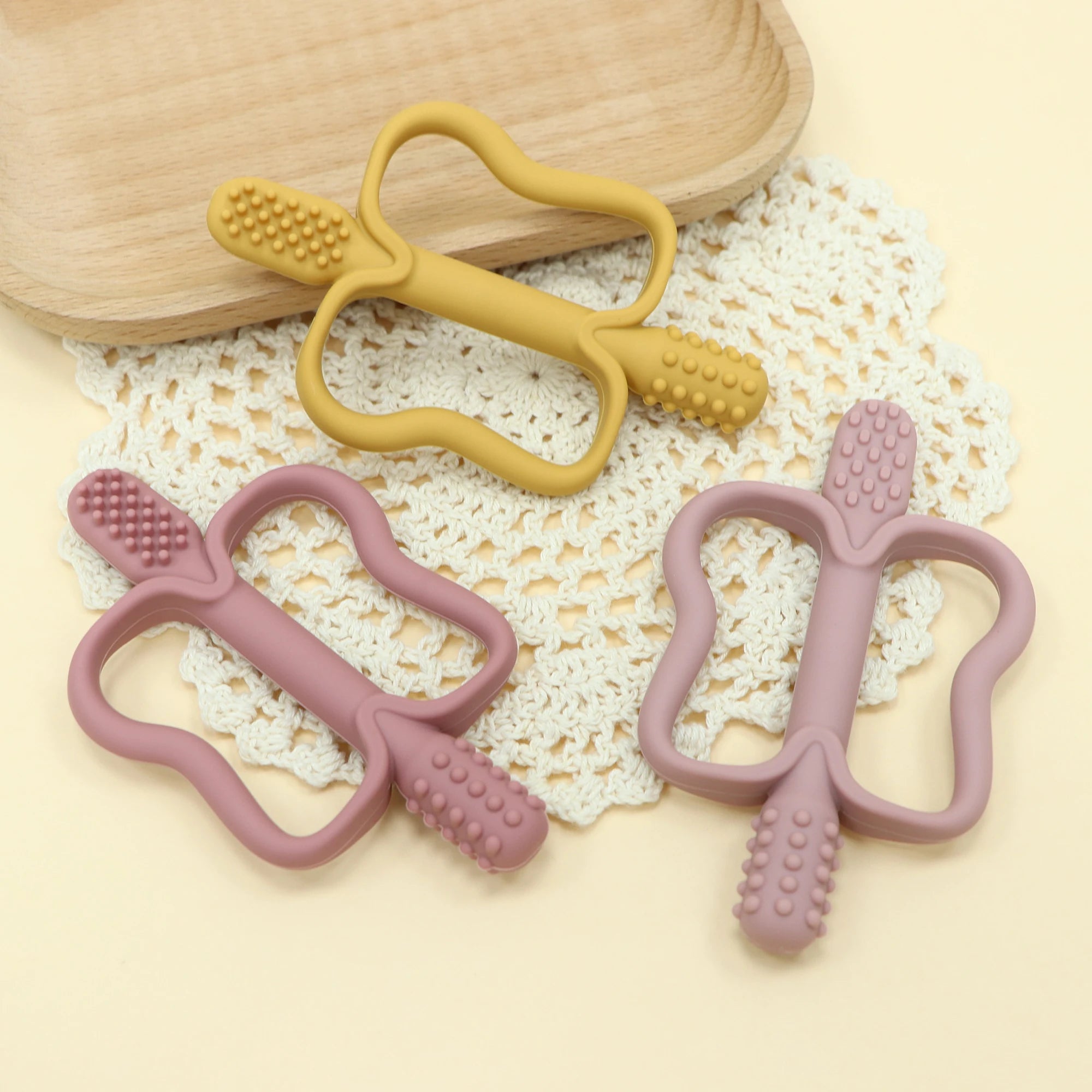 Two yellow and two pink baby teething toys on a textured surface with a wooden bowl in the background.