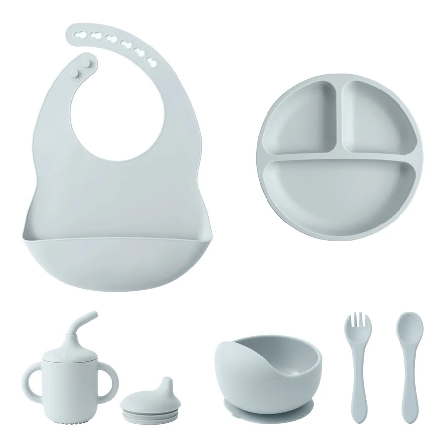 A light gray silicone baby feeding set on a white background, including a bib, divided plate, sippy cup with lid, bowl, and utensils.