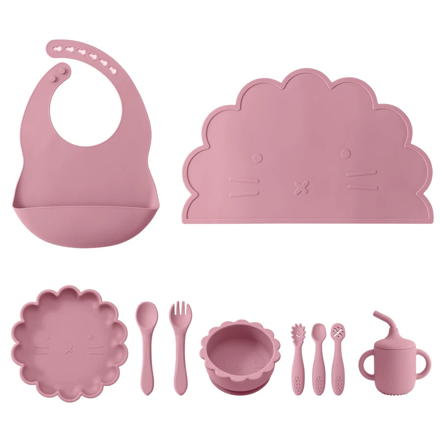 Pink children's dining set with bib, placemat, plate, utensils, and cup on a white background