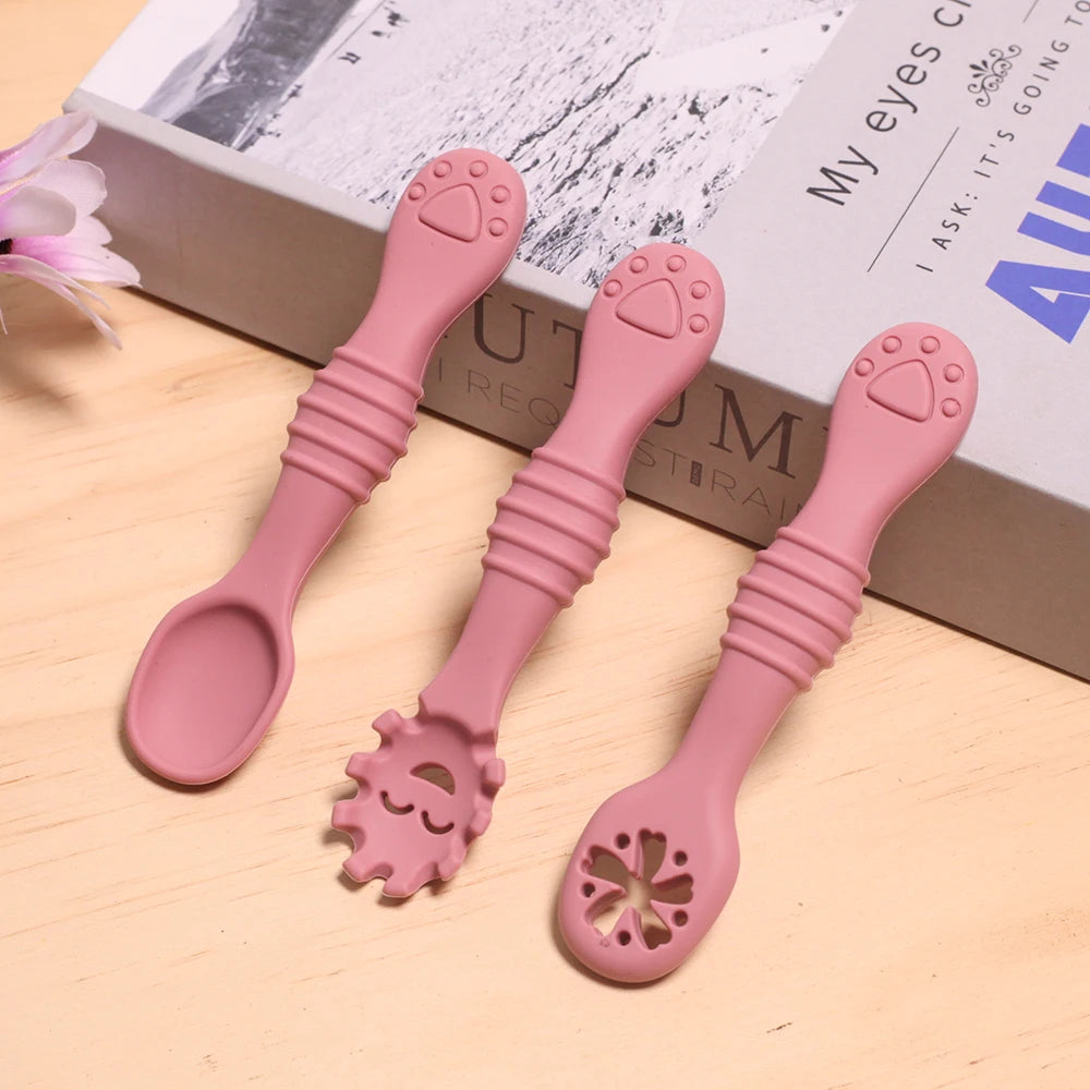 Three pink baby utensils with different designs on a wooden surface.
