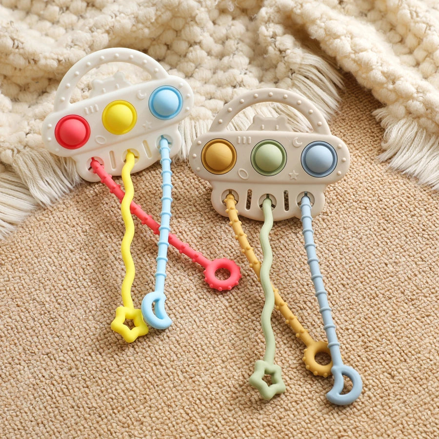 Two baby teething toys with white plastic handles and colorful buttons, resting on a beige textured blanket with a cream knit throw in the background.