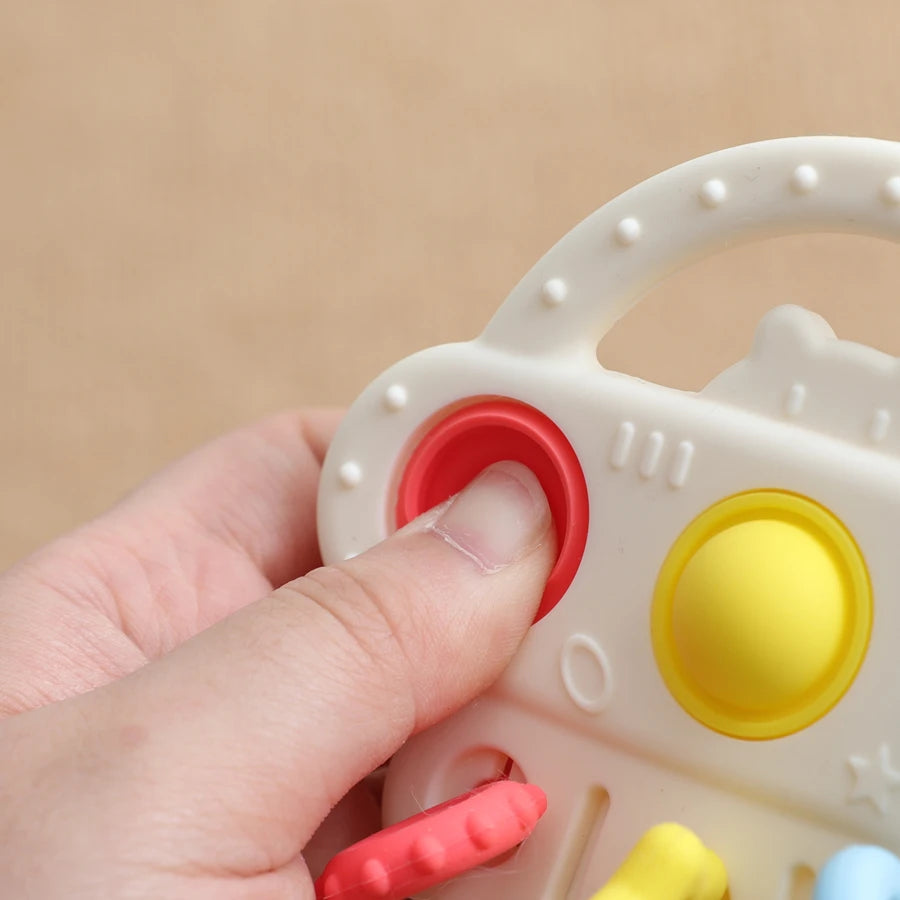 A close-up of a baby teething toy being held by a hand. The toy is primarily off-white plastic with colorful buttons and knobs in red, yellow, and pink, designed for teething.