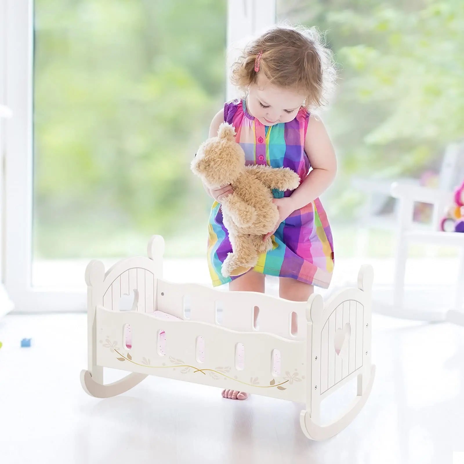 Child in a rainbow dress holding a teddy bear on a white rocking chair with a blurred green outdoor background