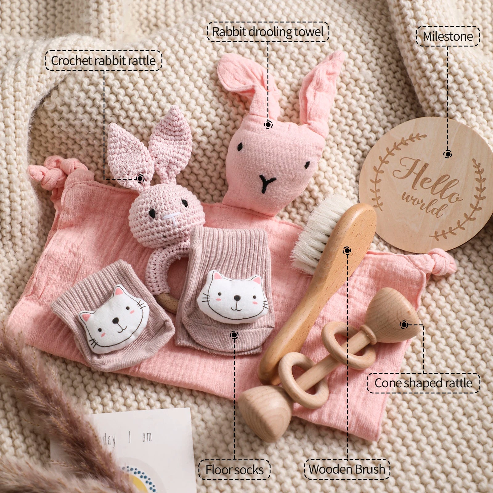 Collection of baby toys and items including a crochet rabbit rattle, drooling towel, milestone card, floor socks, wooden brush, and cone-shaped rattle on a textured surface.