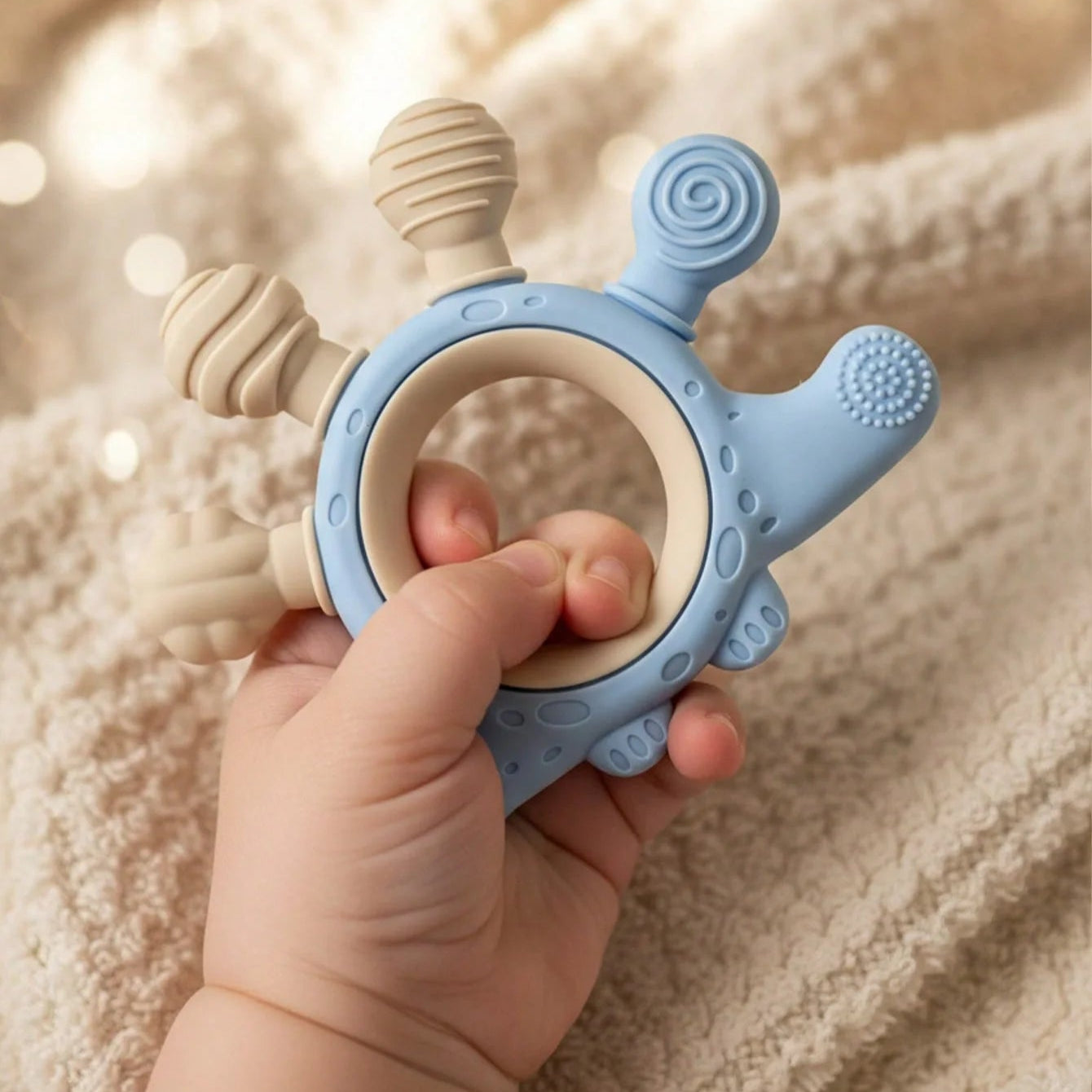 A baby teether toy with a light blue ring and beige textured sensory bumps, held by a baby's hand against a soft beige blanket background.