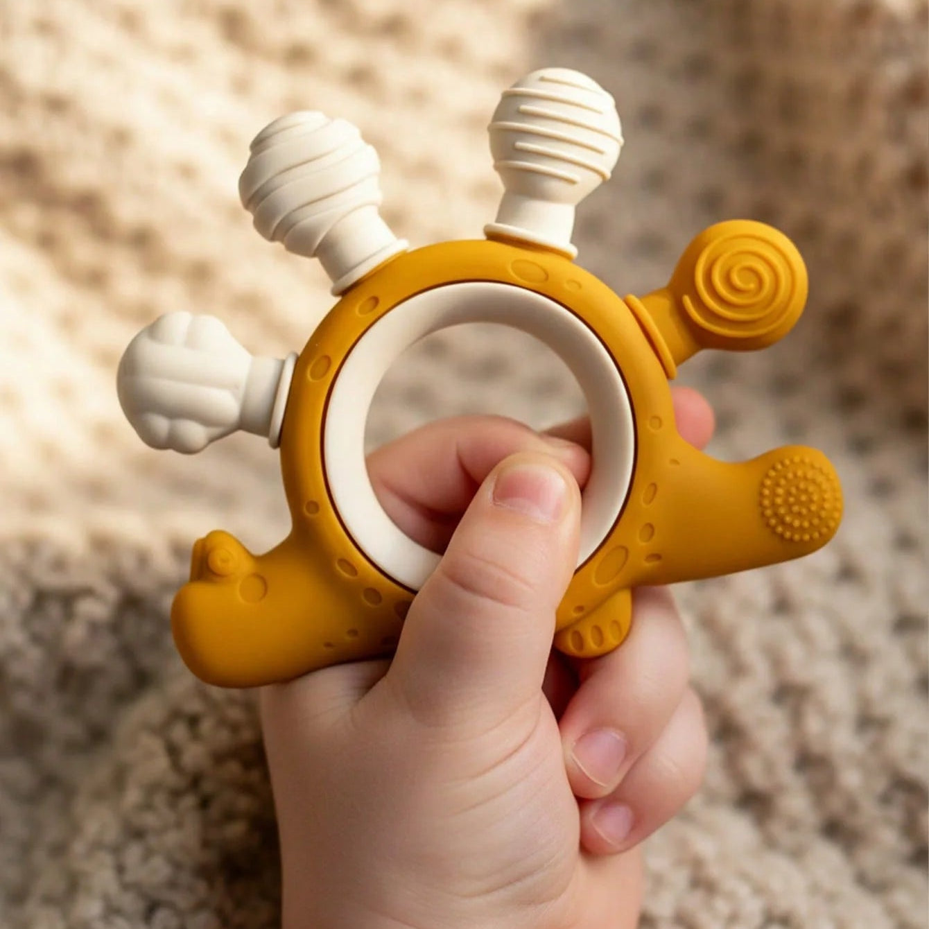 A baby teething ring held in a hand, featuring a mustard yellow dinosaur body with white textured honeycomb, flower, and bubble teeth, set against a soft beige knit blanket background.