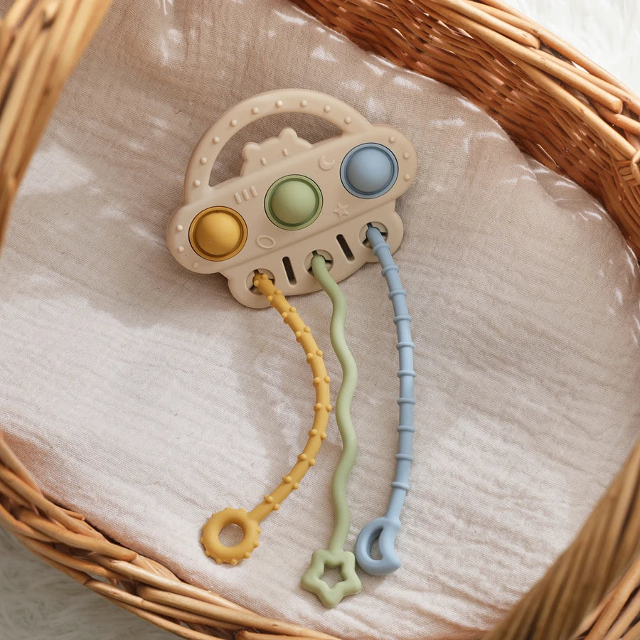 A baby teething toy with a cloud-shaped beige base, three pastel buttons, and three dangling textured rings in yellow, mint green, and light blue, displayed inside a woven wicker basket on soft cream fabric.
