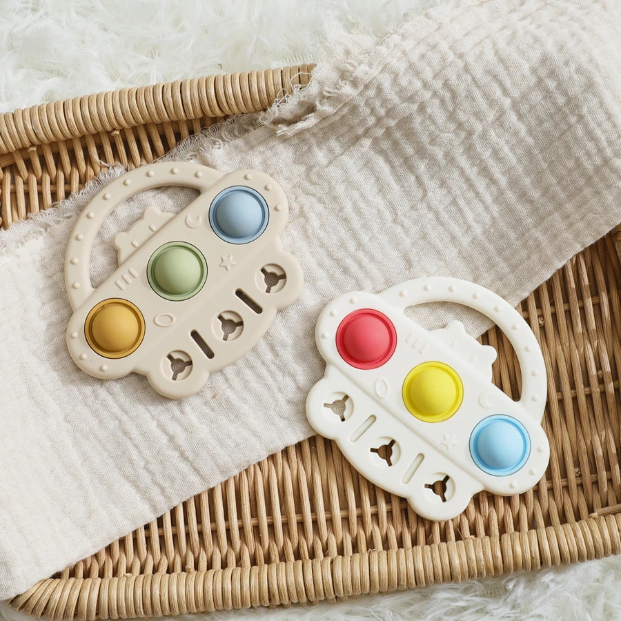 Two baby teething toys shaped like steering wheels, resting on a white textured blanket over a woven wicker tray. One toy is beige with green, yellow, and blue buttons, and the other is white with red, yellow, and blue buttons.
