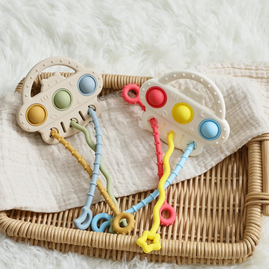 Two baby sensory toy car teething rings with colorful wavy and ring-shaped handles, displayed on a beige muslin cloth inside a woven rattan tray over a white faux fur surface.