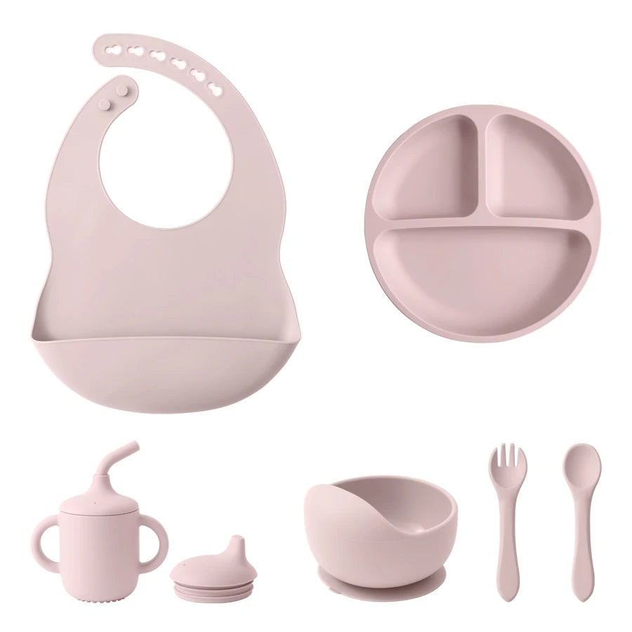 A baby feeding set in a soft dusty pink color, including a silicone bib, a three-compartment divided plate, a handled sippy cup with a spout lid, a small bowl with a suction base, and a fork and spoon.