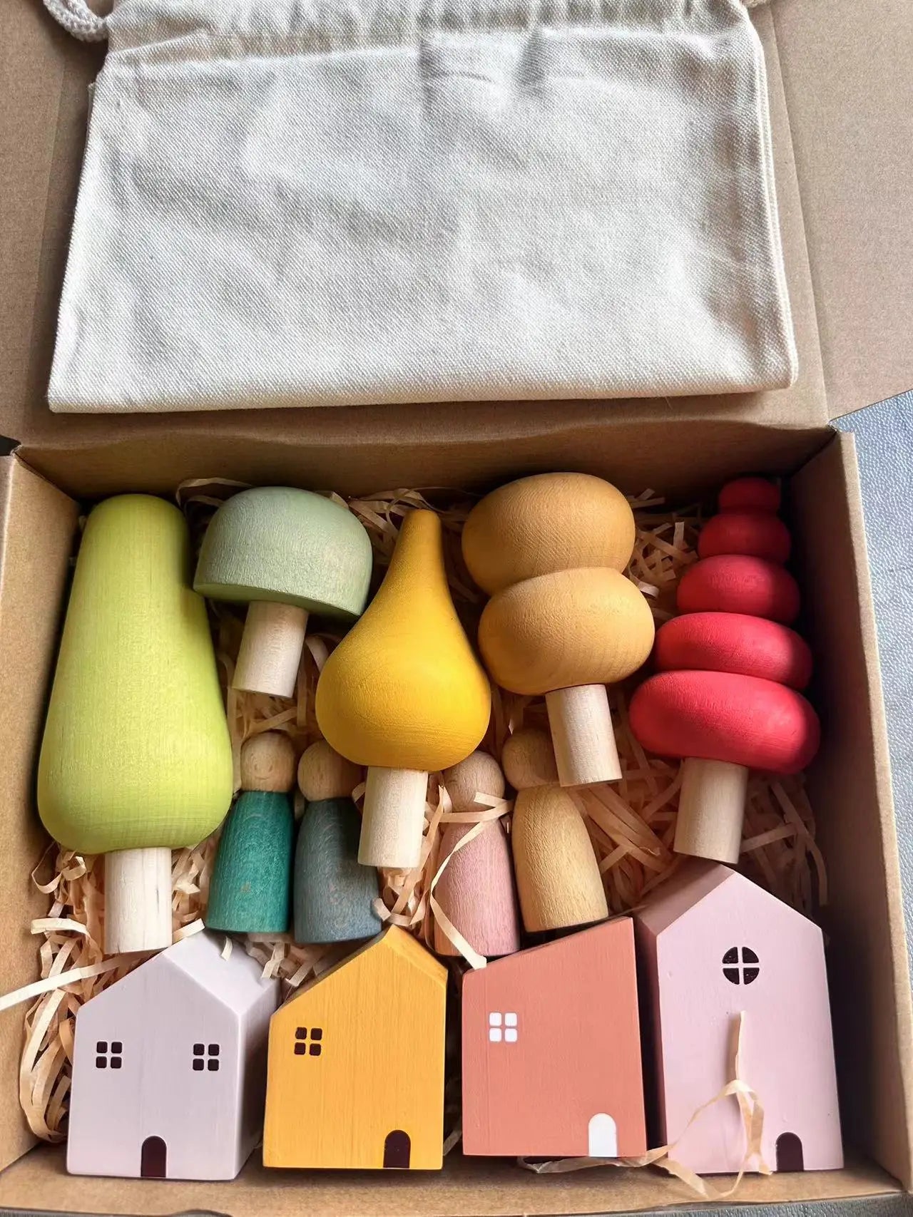 Colorful wooden toy houses and trees in a box with a white cloth inside.