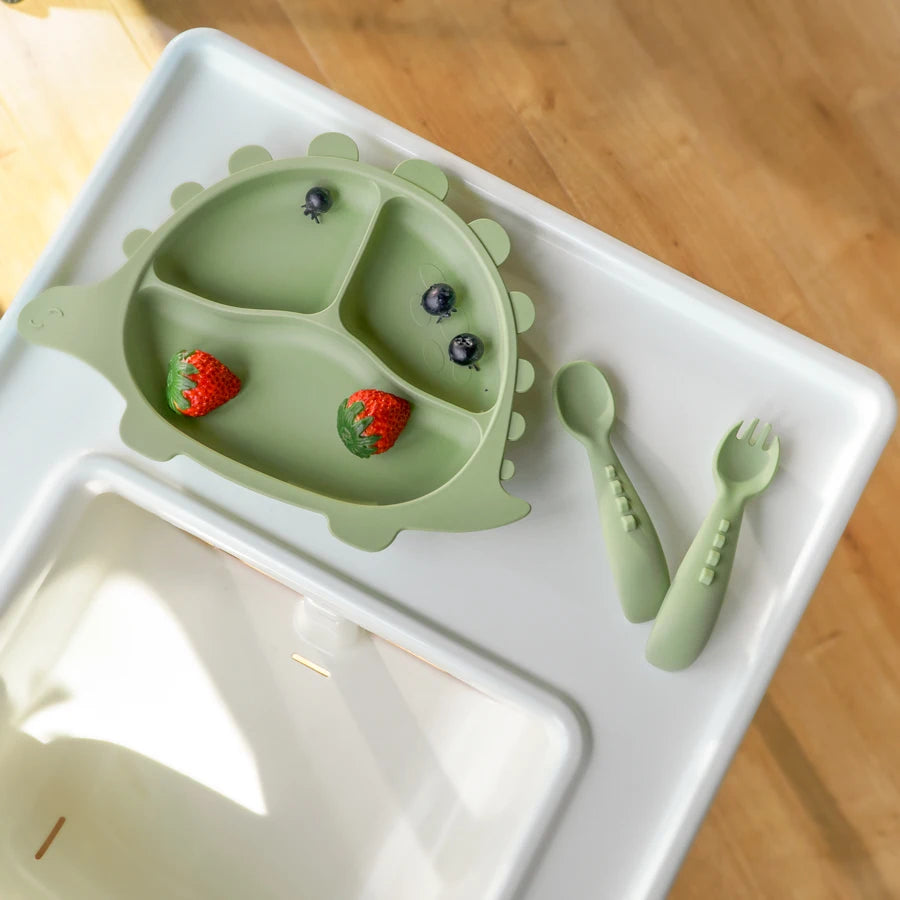 Sage spoon and fork next to a green dinosaur plate with fruit