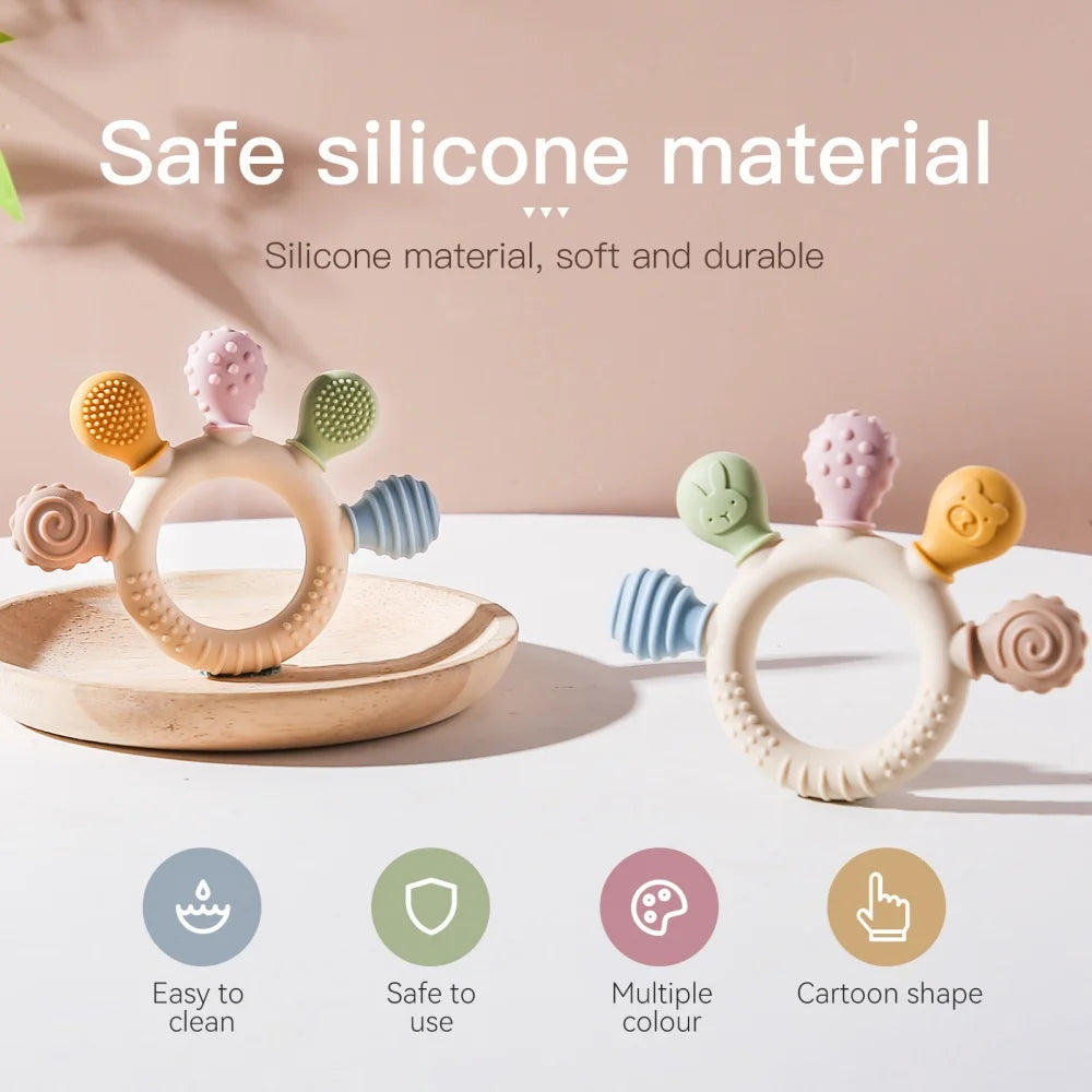 Two pastel-colored silicone baby teething rings with textured and cartoon-shaped silicone attachments, displayed on a light surface with a wooden tray. The image includes text overlays highlighting the safe silicone material and four feature icons at the bottom.