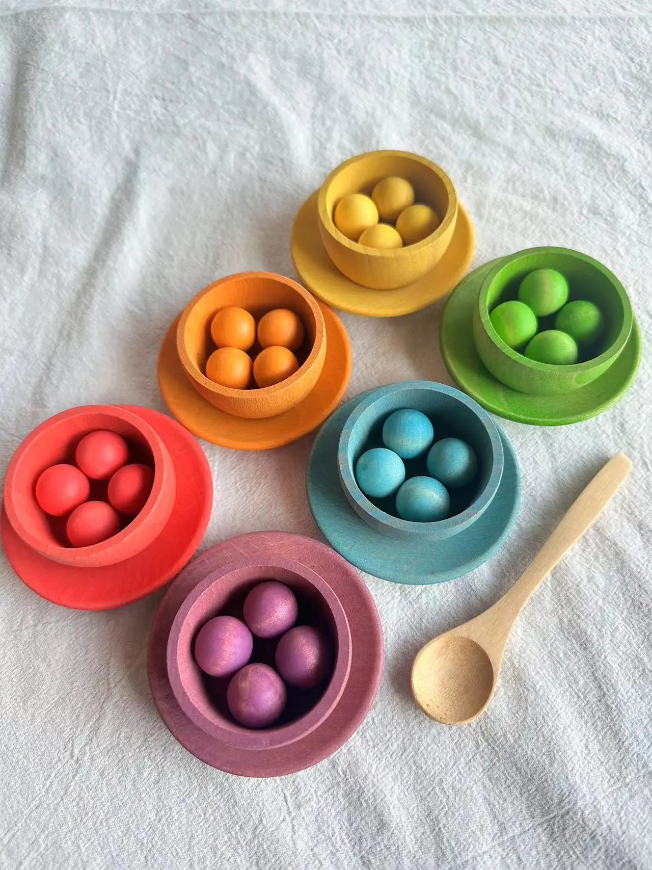 Colorful bowls with spherical objects and a wooden spoon on a white fabric background