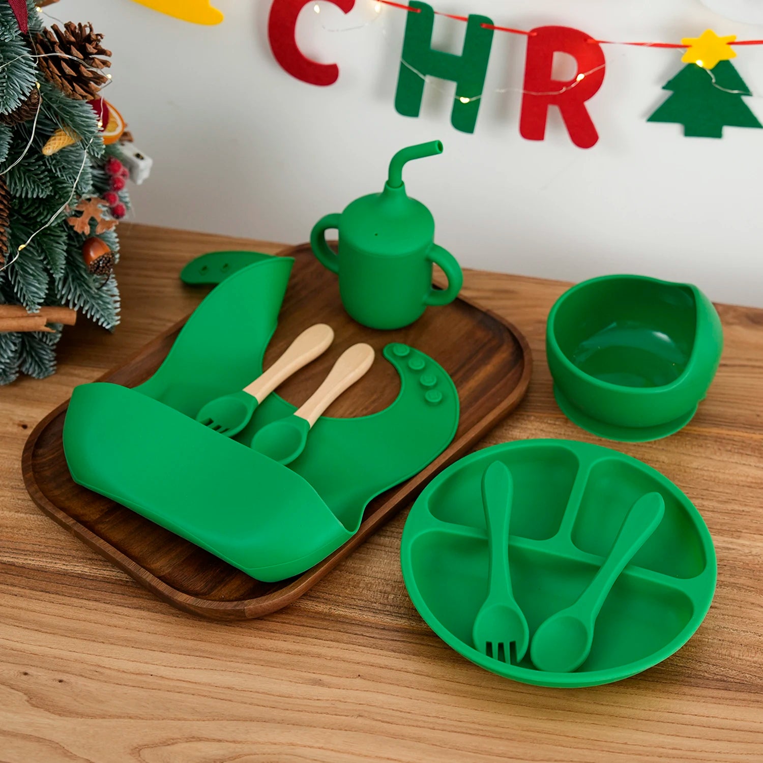 Green silicone bib, plate, bowl, cup and utensils on a wooden tray