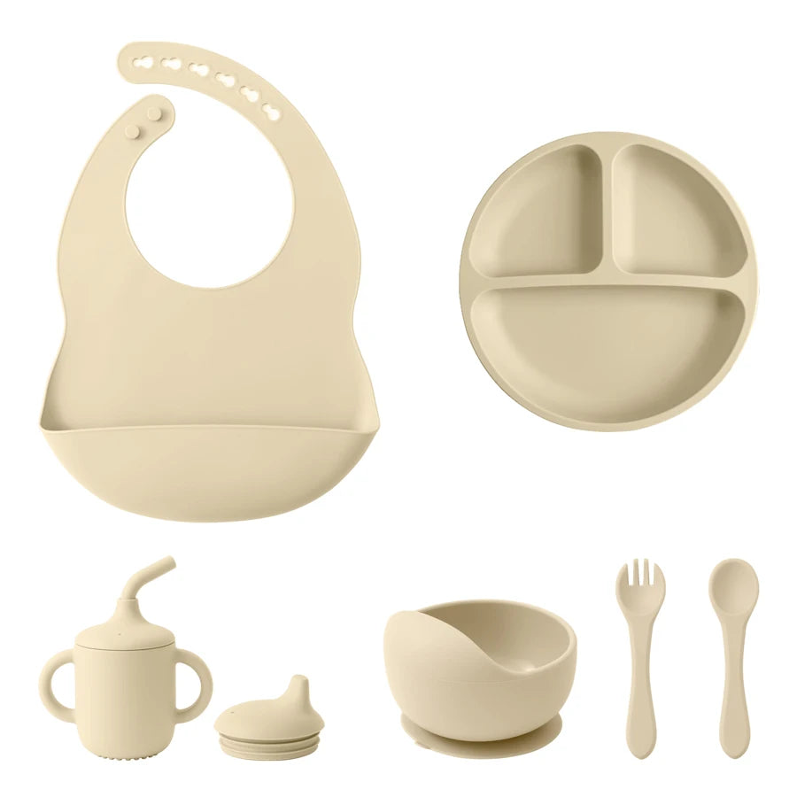 A set of beige baby feeding accessories on a white background, including a silicone bib, a three-compartment divided plate, a sippy cup with spout and cap, a small bowl with a suction base, and a fork and spoon.