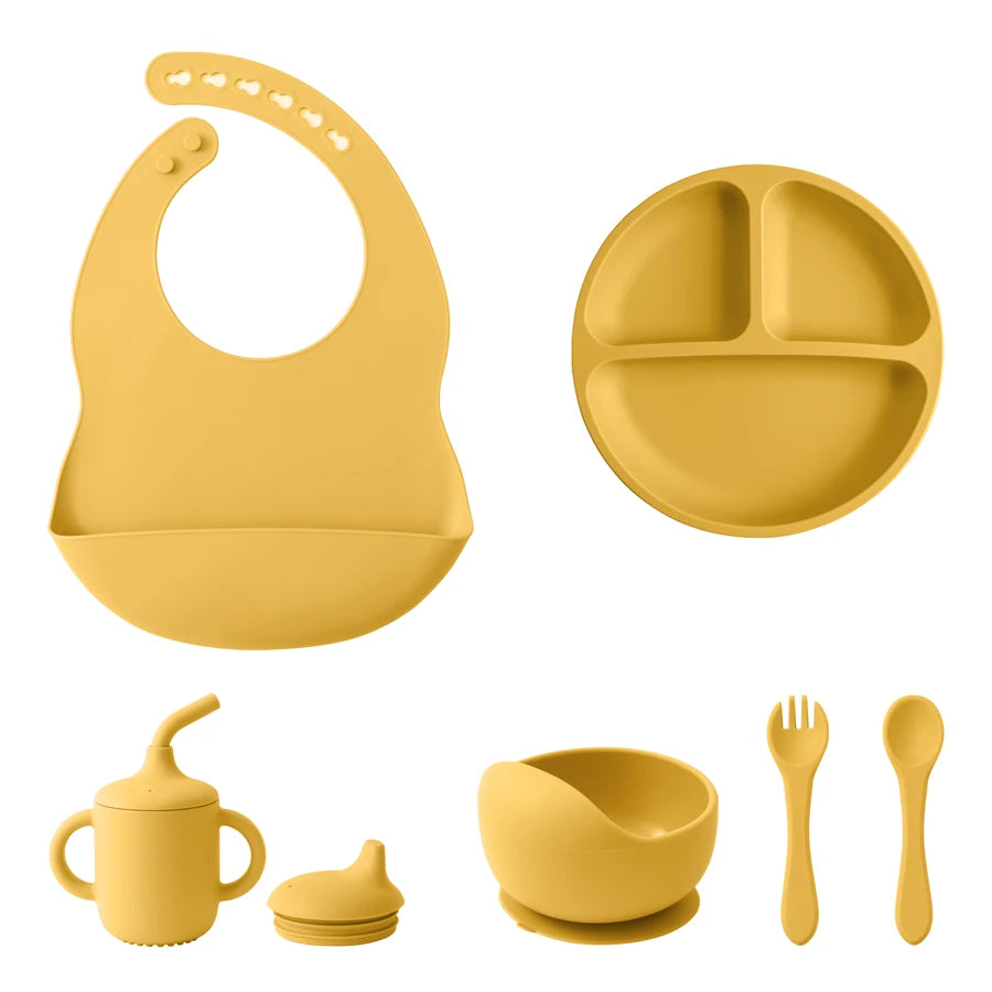 A set of baby feeding accessories in a solid mustard yellow color, including a silicone bib with a suction base, a three-compartment divided plate, a handled sippy cup with a spout lid, a small bowl with a suction base, and a fork and spoon.