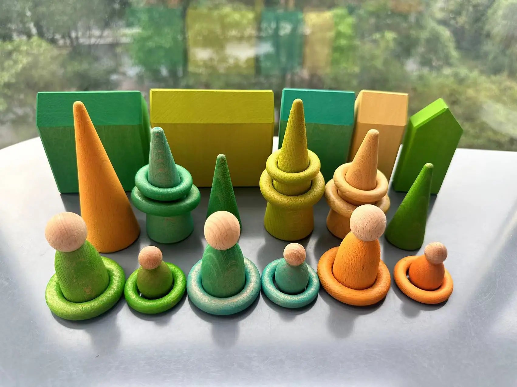 Set of colorful wooden toy figures and blocks on a reflective surface with a blurred outdoor background.