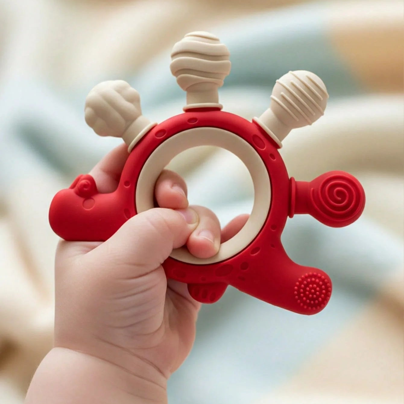 A baby teether toy held in a baby's hand. The toy is made of red and beige silicone with multiple textured sensory bumps and a ring for grasping.