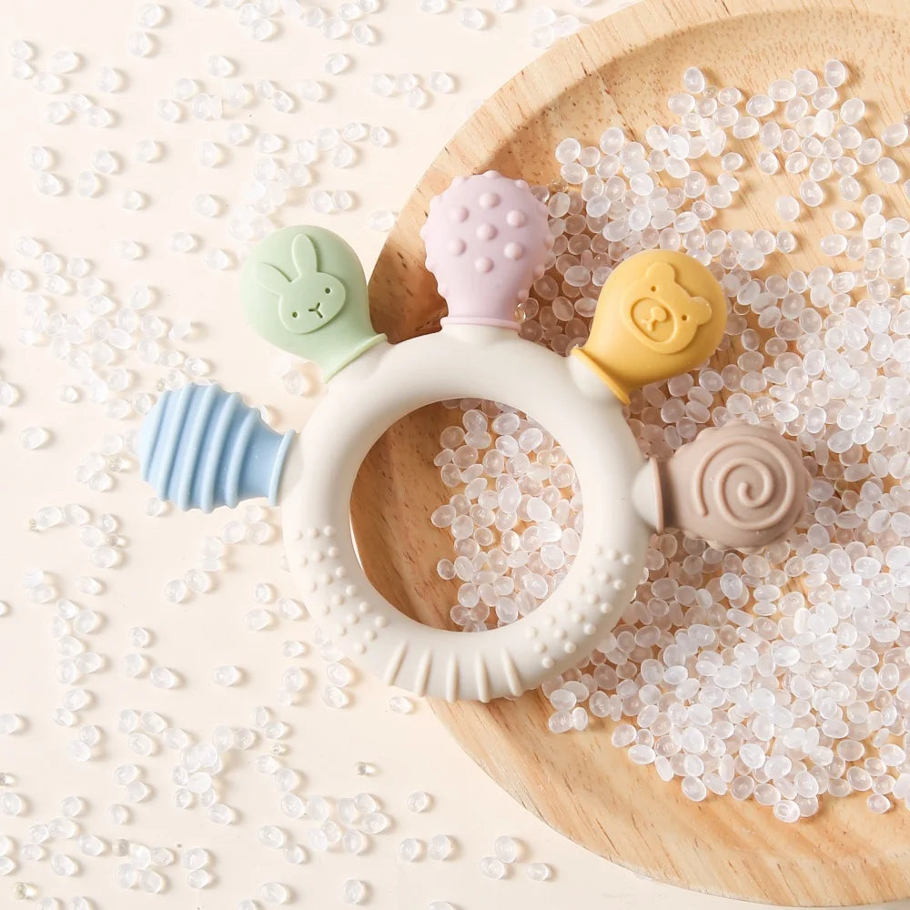A silicone baby teether with a white ring and five pastel-colored textured teethers (green bunny, pink mushroom, yellow bear, blue striped handle, beige spiral) arranged on a wooden plate filled with clear plastic beads on a cream background.