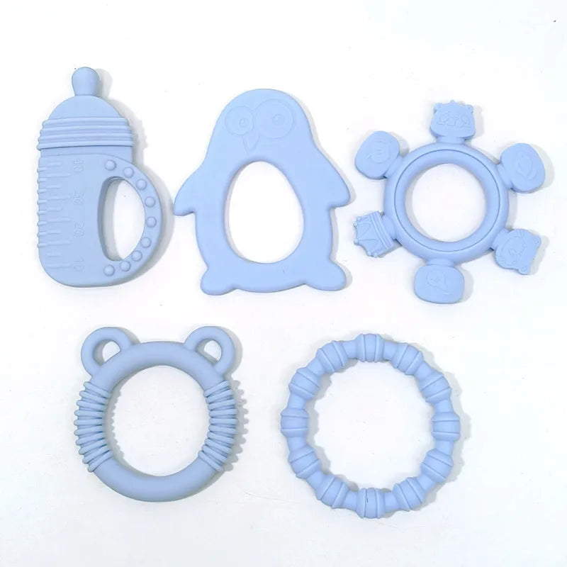 Five light blue silicone baby teething toys arranged on a plain white background, including a bottle-shaped teether, an owl teether, a flower ring teether, a bear-tear ring teether, and a textured circular teether.
