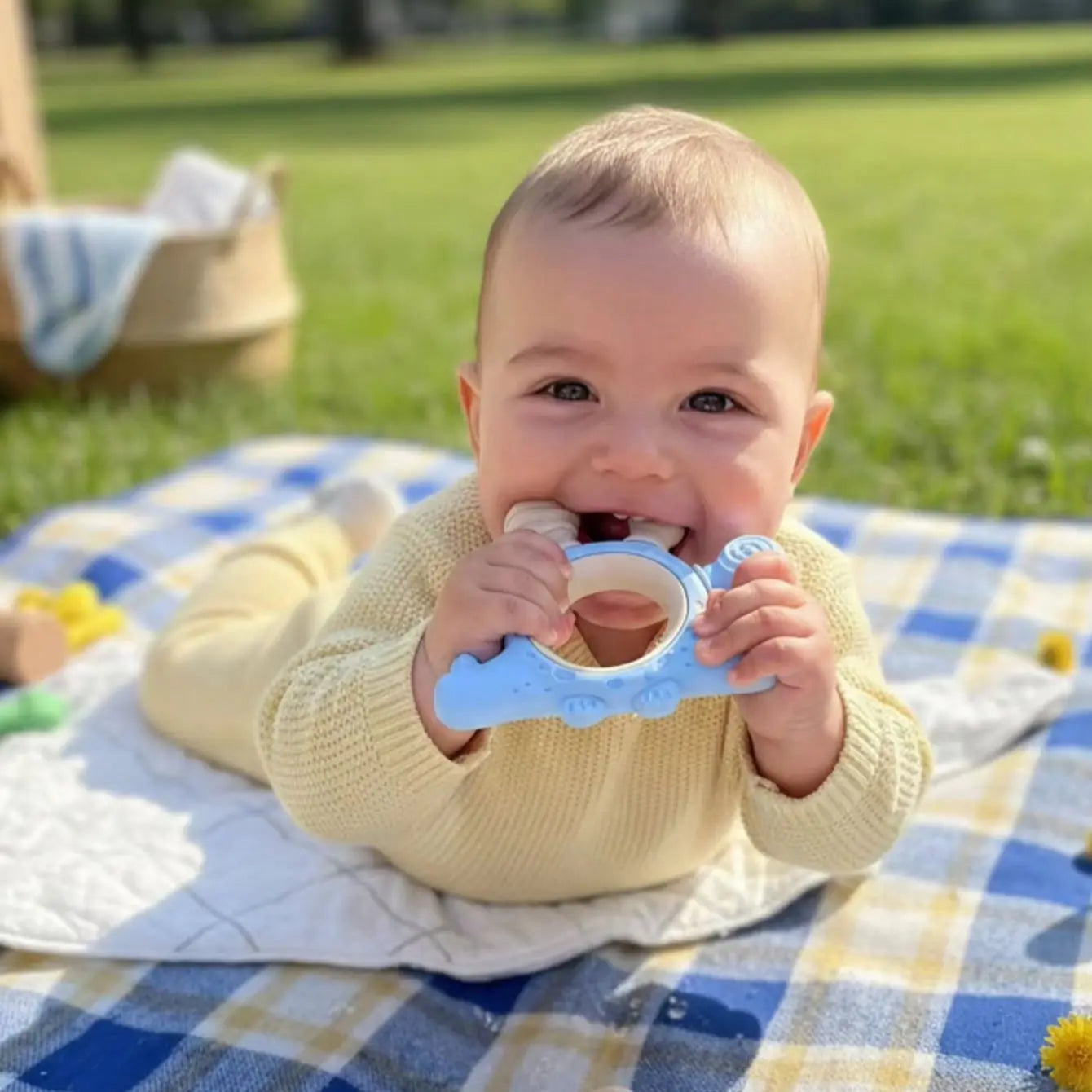 A baby wearing a cream knit sweater lies on a blue and white check blanket on a grassy lawn, holding a light blue silicone teether.
