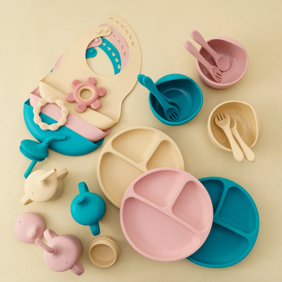 A pastel baby feeding set arranged on a warm cream background, including divided plates, bowls, utensils, a bib, tees, and small accessories in pink, blue, and beige.