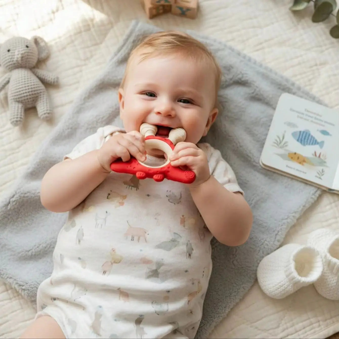 A baby lying on a light gray blanket, biting a red silicone teether with wooden teethers. The baby is wearing a white onesie with a soft animal print.