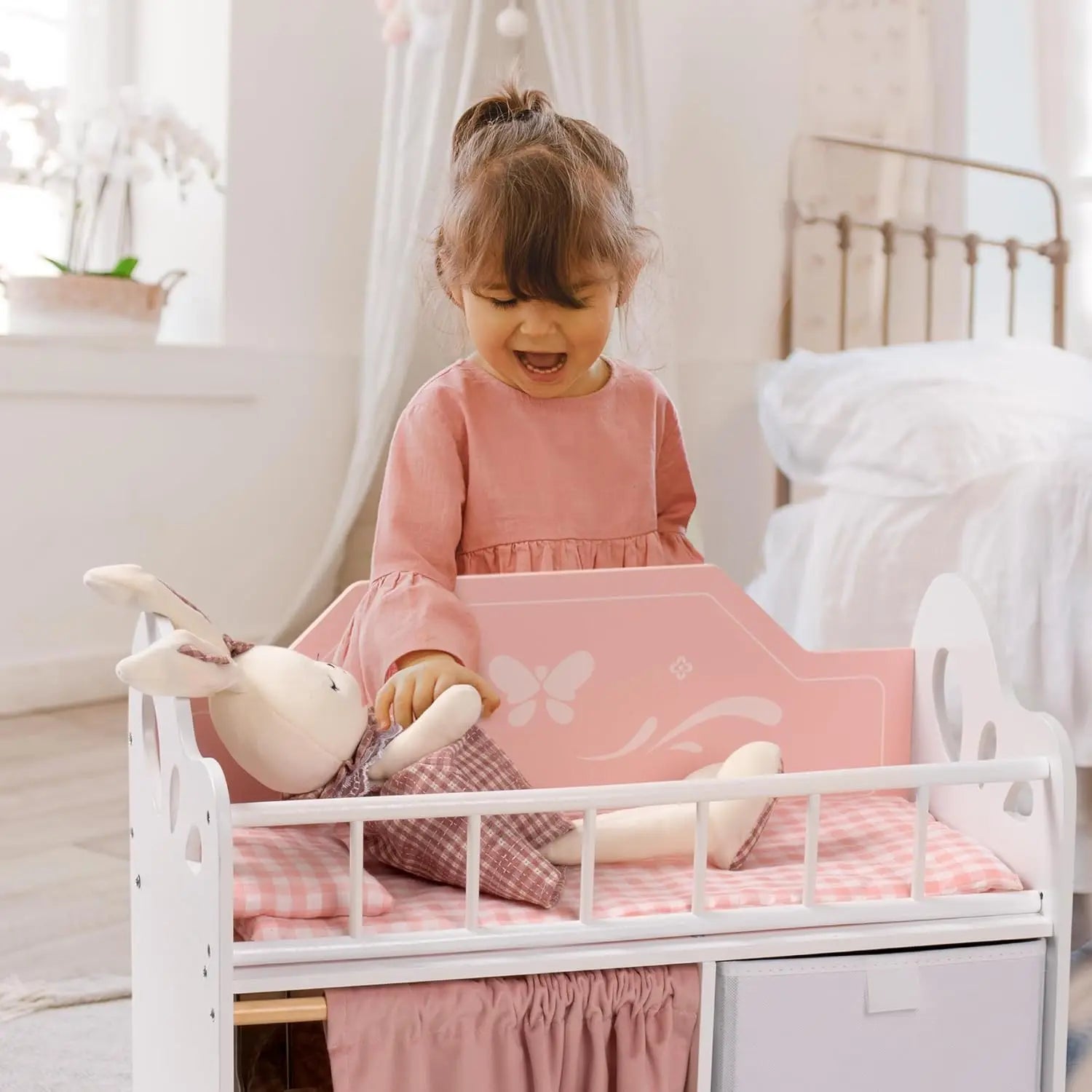 Child in a pink dress sitting on a white crib with pink bedding and toys.