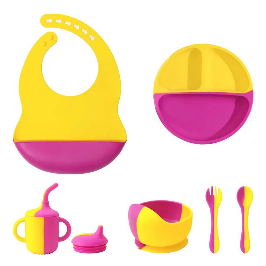 A colorful baby feeding set on a plain white background, including a silicone bib, a divided plate, a handled sippy cup with lid, a suction bowl, and a fork and spoon.