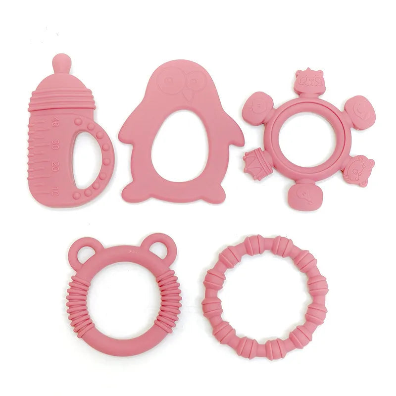 Five pink silicone baby teething toys arranged on a plain white background, including a bottle-shaped toy, a penguin-shaped toy, a flower-shaped ring toy, a bear-ear ring toy, and a textured circular ring toy.